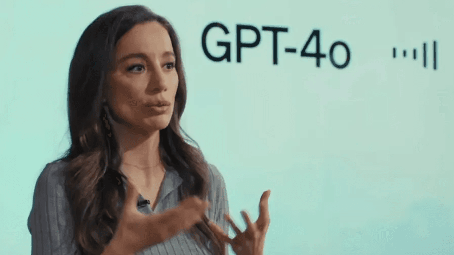 Former OpenAI CTO Mira Murati presenting on stage during the GPT-4o launch event, gesturing while speaking in front of a screen displaying the GPT-4o logo and audio waveform visualization.