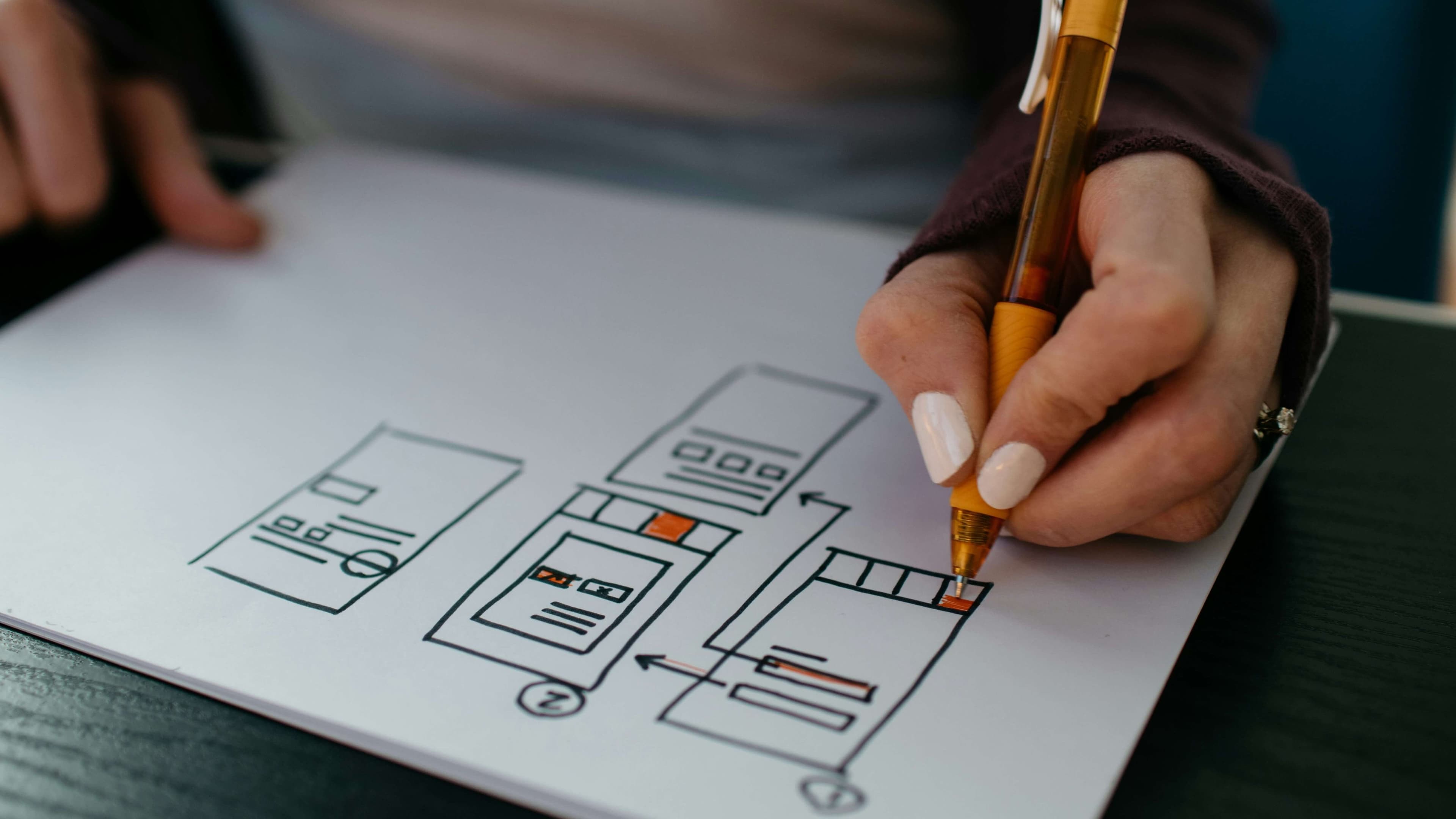 A hand holding an orange pen sketches wireframe mockups on white paper, showing multiple mobile screen layouts connected by arrows to indicate user flow. The wireframes include rough rectangles representing screens with placeholder elements for headers, content blocks, buttons, and navigation icons, with some elements highlighted in orange and red to denote interactive components or calls to action. The sketch illustrates a multi-step interface flow typical of login or onboarding design planning.